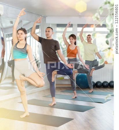 Girl in sportswear exercising with group multinational people at yoga studio, standing in warrior one leg pose on fitness mat Girl in sportswear exercising with group multinational people at yoga studio, standing in warrior one leg pose on fitness mat 122556801