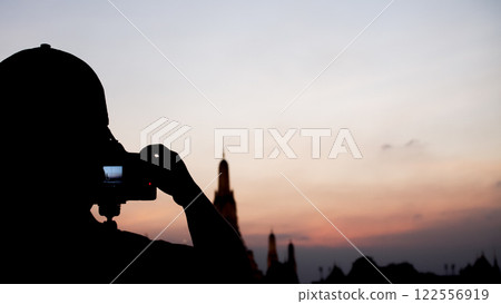 a silhouetted photographer taking a picture of a temple at sunset. The warm sky contrasts with the dark foreground, emphasizing the serene atmosphere. 122556919