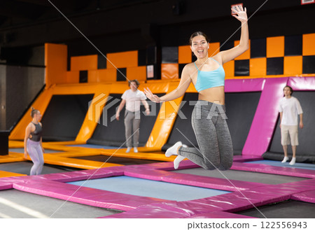 Excited young woman in blue top and gray leggings having great time while jumping on colorful trampoline in game club Excited young woman in blue top and gray leggings having great time while jumping on colorful trampoline in game club 122556943