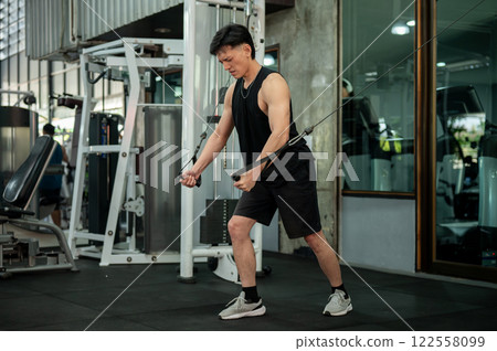 A serious, muscular Asian male athlete is using a cable machine in the gym. 122558099