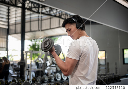 A rear view of a muscular and strong Asian man is working out with dumbbells in the gym. A rear view of a muscular and strong Asian man is working out with dumbbells in the gym. 122558110