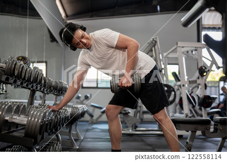 A handsome Asian man is working out with a dumbbell in front of the rack in the gym. A handsome Asian man is working out with a dumbbell in front of the rack in the gym. 122558114