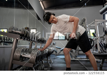 A muscular and handsome Asian man is working out with a dumbbell in front of the rack in the gym. A muscular and handsome Asian man is working out with a dumbbell in front of the rack in the gym. 122558115