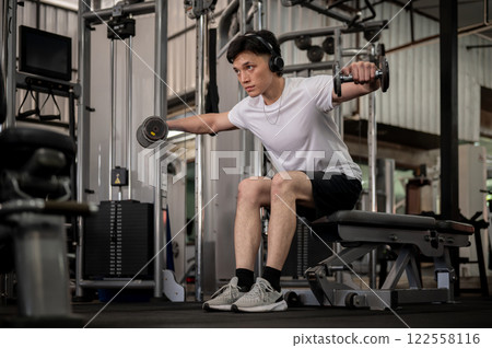 A determined Asian man sits on a fitness bench, working out with dumbbells in the gym. A determined Asian man sits on a fitness bench, working out with dumbbells in the gym. 122558116