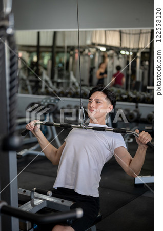 A strong and determined Asian man is working out with a cable machine to train his chest muscles. 122558120