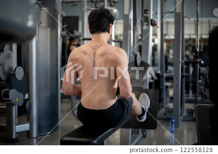 A back view of a strong, shirtless Asian man is exercising with a multi station machine in a gym. 122558128