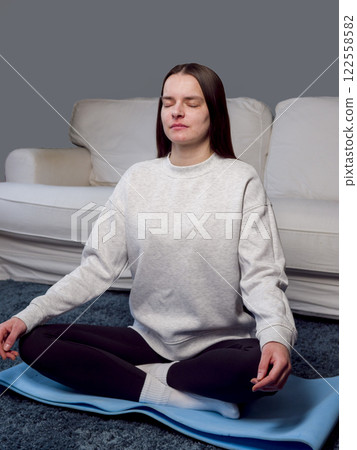 Serene woman meditating in lotus position on yoga mat, eyes closed, finding calm within minimalist living room space, practicing mindfulness vertical photo Serene woman meditating in lotus position on yoga mat, eyes closed, finding calm within minimalist living room space, practicing mindfulness vertical photo 122558582
