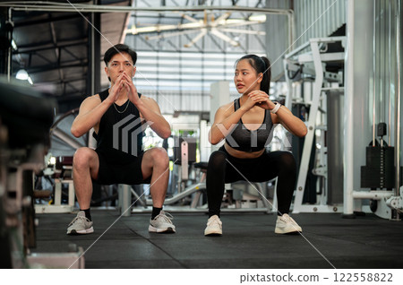 A sporty Asian couple is training together in the gym, performing squats and bodyweight exercises. 122558822