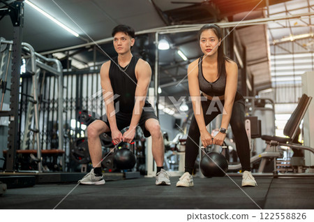 A sporty and determined Asian male and female are performing squats with a kettlebell in the gym. 122558826