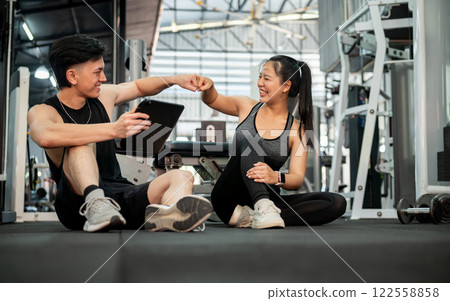 A cheerful Asian male fitness trainer and his client are in the gym, giving each other a fist bump. A cheerful Asian male fitness trainer and his client are in the gym, giving each other a fist bump. 122558858