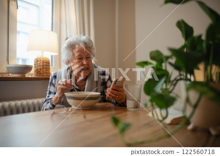 Elderly woman using smartphone while sitting in kitchen and eating soup. 122560718