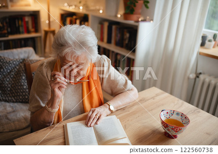 Lovely elderly lady reading a book, sitting at the kitchen table and sipping tea. 122560738