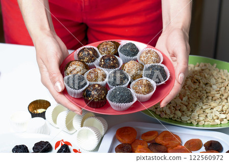 A woman holds in her hands a plate with dried fruit sweets. From prunes, dates and coconut. With a sprinkle of black sesame powder. A woman holds in her hands a plate with dried fruit sweets. From prunes, dates and coconut. With a sprinkle of black sesame powder. 122560972