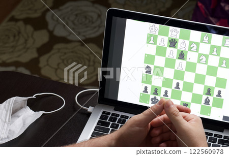 A man solves a chess problem online. Chess board with figures on a laptop screen. Leisure in self-isolation, in the twilight. International chess day. 122560978