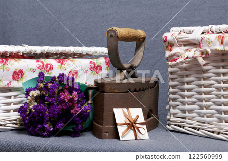 Old iron, heated by hot coals. Located on gray fabric. Nearby are wicker baskets, a bouquet of dried flowers and a greeting card. 122560999