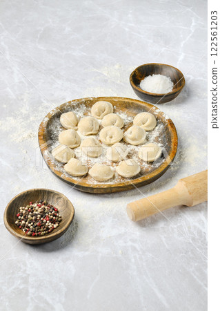 Freshly made dumplings arranged on a wooden plate with spices and rolling pin on a marble surface 122561203