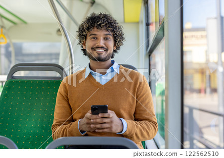 A man uses an app on his smartphone while riding public transport, smiling and looking at the camera with the phone in his hands. A man uses an app on his smartphone while riding public transport, smiling and looking at the camera with the phone in his hands. 122562510