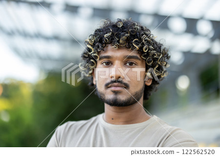 Close-up portrait of a young calm and serious man, looking at the camera, walking outside in the park. Close-up portrait of a young calm and serious man, looking at the camera, walking outside in the park. 122562520