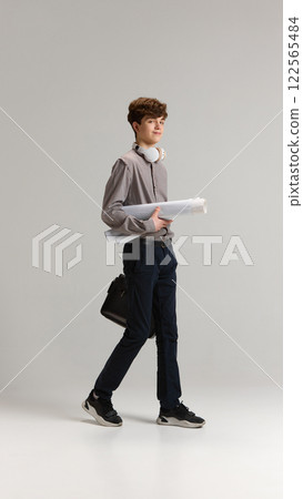 Full-length portrait of teen boy, student in shirt and jeans, walking with briefcase and papers, project against grey studio background Full-length portrait of teen boy, student in shirt and jeans, walking with briefcase and papers, project against grey studio background 122565484