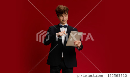Teen boy, student in formal wear with bow tie interacting with tablet against red studio background. Online communication, remote lessons Teen boy, student in formal wear with bow tie interacting with tablet against red studio background. Online communication, remote lessons 122565510