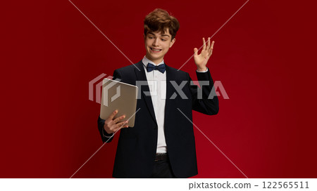 Portrait of smiling teen boy, student on formal wear with bow tie holding tablet and waving towards screen against red studio background Portrait of smiling teen boy, student on formal wear with bow tie holding tablet and waving towards screen against red studio background 122565511