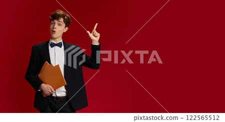Portrait of young man, student on formal wear with bow tie standing with papers and raising finger up, showing gesture of idea against red studio background Portrait of young man, student on formal wear with bow tie standing with papers and raising finger up, showing gesture of idea against red studio background 122565512