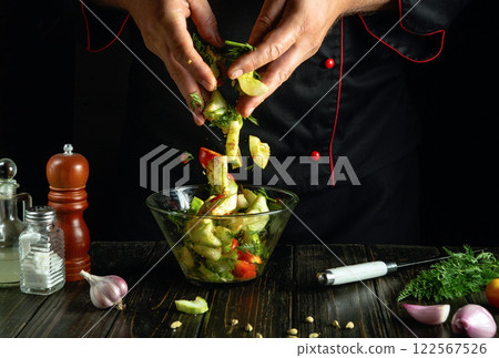 With expert hands, a chef tosses a mix of colorful vegetables into a clear bowl, creating a delicious salad. Aromatic herbs fill the air, enhancing the lively kitchen atmosphere 122567526