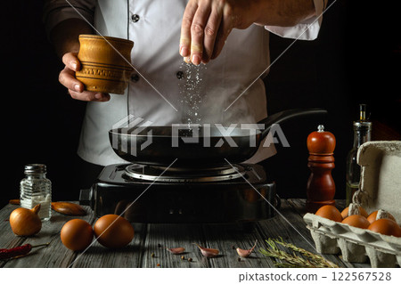 A skilled chef prepares a meal in a cozy kitchen, delicately adding salt to a sizzling pan filled with eggs, surrounded by fresh ingredients. The warm ambiance combines tradition and culinary art 122567528