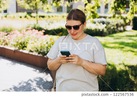 Happy woman in sunglasses chatting on phone and relaxing outdoors. Urban people concept. Smiling 30s woman text messaging on smart phone at sunny day. Happy woman in sunglasses chatting on phone and relaxing outdoors. Urban people concept. Smiling 30s woman text messaging on smart phone at sunny day. 122567536