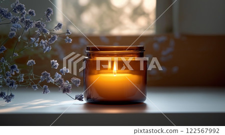 Amber jar candle glowing softly on a windowsill, surrounded by lavender flowers in sunlight 122567992