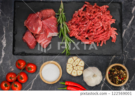 Finely chopped red minced meat and pieces of beef lie on a black cutting board, separated by sprigs of rosemary on a dark background on top, vegetables and seasonings at the bottom. The concept of 122569001