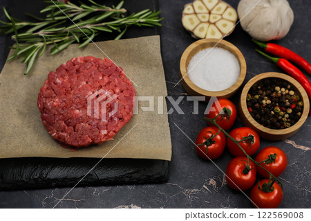 Close-up on the left of a raw beef round patty lying on paper next to seasonings, red hot peppers, garlic, rosemary and tomatoes on a branch on a dark background. High quality photo 122569008