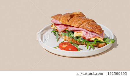 Croissant sandwich with ham, cheese and arugula on a plate on a yellow background with a shadow. Croissant sandwich with ham, cheese and arugula on a plate on a yellow background with a shadow. 122569395