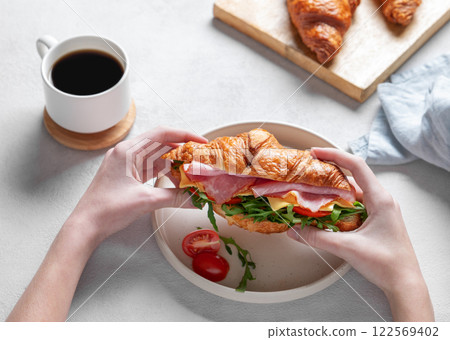 Hands holding fresh croissant sandwich with ham, cheese, tomato  and arugula on a plate  122569402
