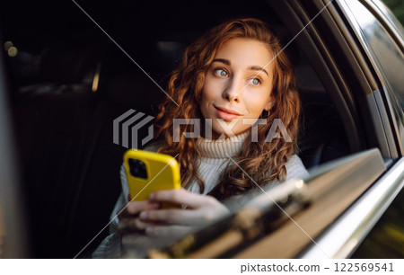 Positive young woman in casual clothes sits in the back seat of a car with a mobile phone. Positive young woman in casual clothes sits in the back seat of a car with a mobile phone. 122569541
