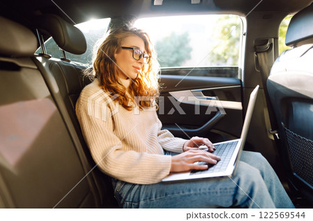 Young woman in glasses with a laptop in the back seat of a car. Freelance, business concept. Young woman in glasses with a laptop in the back seat of a car. Freelance, business concept. 122569544