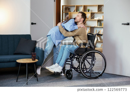 Young man in wheelchair and his wife hugging, standing in living room at home 122569773