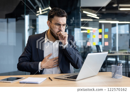 Sick man coughing and having a cold at workplace. Businessman working with laptop at workplace inside office. Sick man coughing and having a cold at workplace. Businessman working with laptop at workplace inside office. 122570162