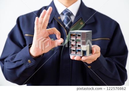 一名身穿深藍色工作服的中年男子手持公寓大樓模型，比畫著圓圈手勢 122570862