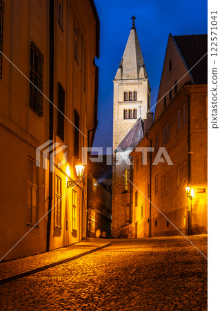 An evening view of the Bell Tower of St. George's Basilica from Jirska Street, illuminated by street lamps and showcasing the historic architecture of Prague. 122571041