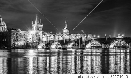 The historic Charles Bridge elegantly spans the Vltava River, illuminated under a night sky in Prague. The serene water reflects the bridge's arches and nearby architecture. 122571043