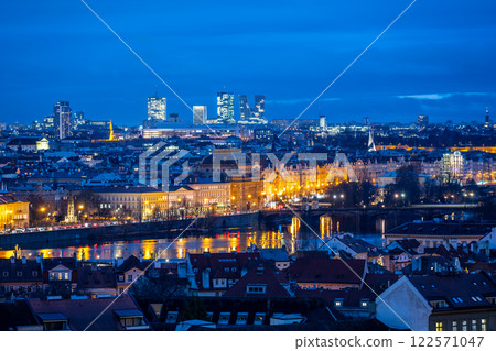 The National Theatre stands illuminated against the evening sky in Prague, with modern buildings of Pankrac rising in the background. Reflections shimmer in the water. 122571047