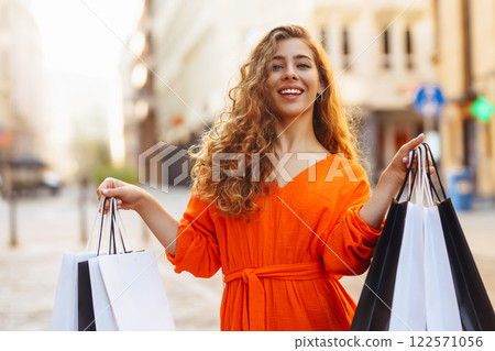 Cheerful young woman with shopping bags walking down city street. Purchases, black friday, discounts Cheerful young woman with shopping bags walking down city street. Purchases, black friday, discounts 122571056