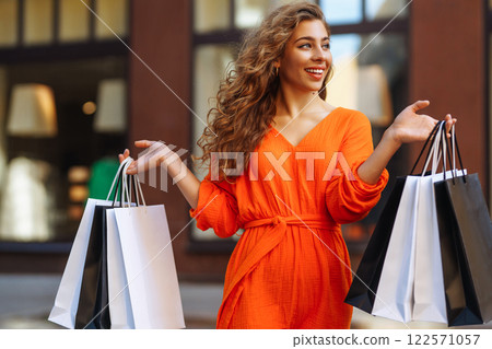 Cheerful young woman with shopping bags walking down city street. Purchases, black friday, discounts 122571057