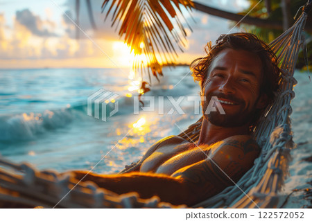 Cheerful man lying comfortably on a hammock strung between two palm trees on a deserted beach at sunset, enjoying the serene tropical surroundings 122572052