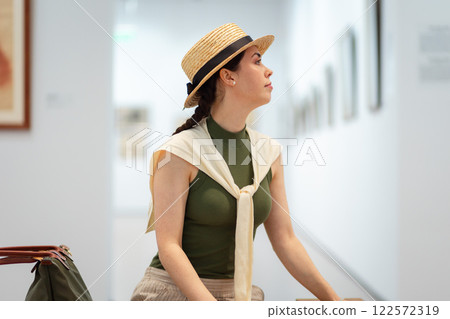 Mid shot of young pretty Caucasian woman in a straw hat and elegant clothes visits a contemporary art gallery. Side view. Museum exhibition Mid shot of young pretty Caucasian woman in a straw hat and elegant clothes visits a contemporary art gallery. Side view. Museum exhibition 122572319