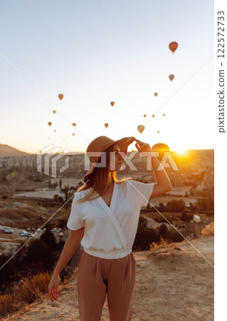 Woman stands on mountain with flying air balloons on background. Tourist Turkish region Cappadocia. Woman stands on mountain with flying air balloons on background. Tourist Turkish region Cappadocia. 122572733