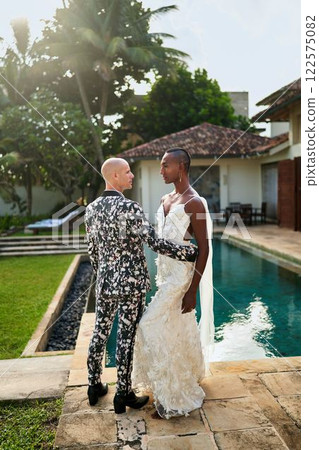 Interracial queer couple wedding at villa. Ambiguous black bride in white dress, bold groom in patterned suit. LGBTQ-friendly tropical ceremony, elegant, vibrant love celebration. 122575082
