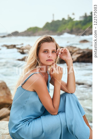 Blonde woman in blue dress sits on rocky ocean shore. She thoughtful with hand on face. Fashionable and serene moment. Coastal vibes. High-end female beauty, fashion scene. 122575083