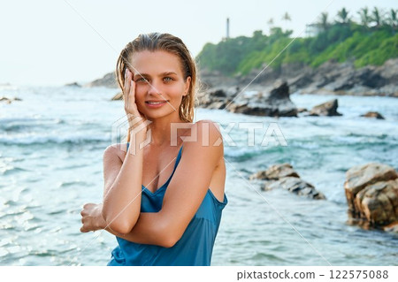 Young woman in blue swimsuit smiles by ocean wet hair, relaxed pose. Tropical rocky coastline in background. Sunlight enhances serene beach atmosphere. Natural beauty embraced by sea breeze. 122575088